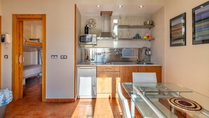 A compact kitchen with a microwave, stove, and sink, a glass dining table, and a small bed in the adjacent room. - Apartment 'Diseño A Pie De Pista' with Mountain View and Wi-Fi (Monachil)