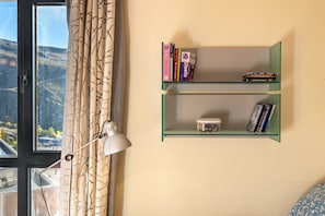 A room with a window, a lamp, and two green shelves with books and a container.