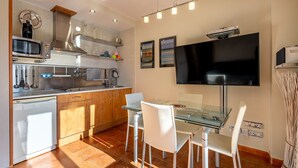 A modern kitchen with a glass dining table, white chairs, a built-in microwave, and a flat-screen TV. - Apartment 'Diseño A Pie De Pista' with Mountain View and Wi-Fi (Monachil)