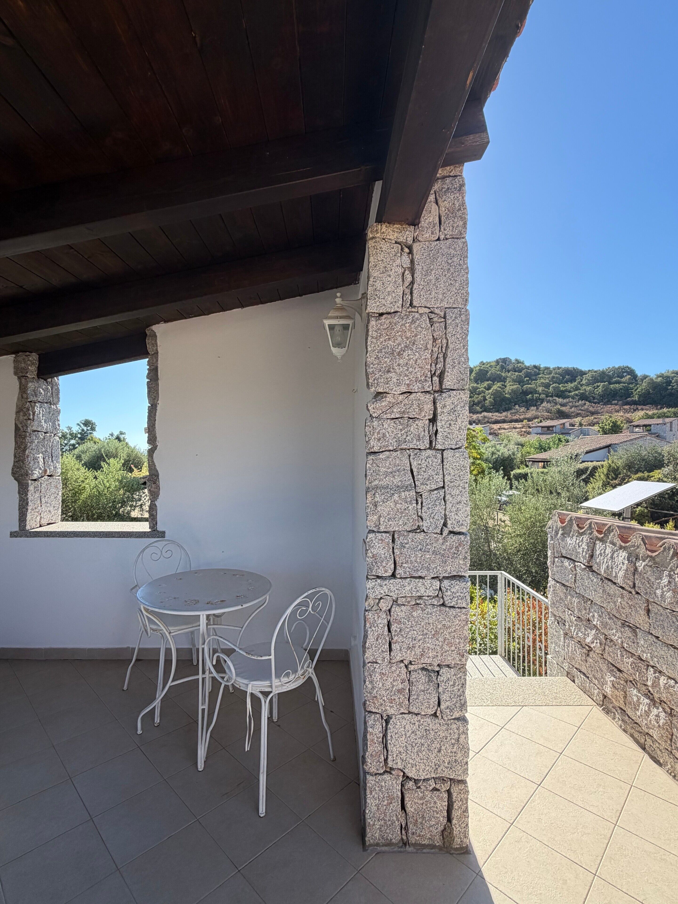 A covered outdoor seating area with a stone wall, a table with chairs, and a view of a landscape with houses and trees.