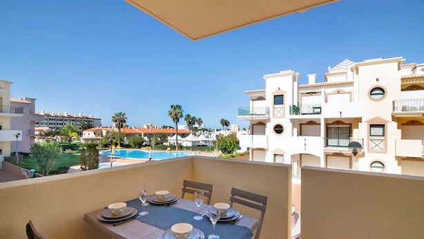 A balcony with a table set for dining, overlooking a swimming pool and buildings.