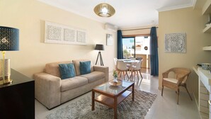 A living room with a sofa, coffee table, and wicker chair, a dining area with chairs and a table, and a balcony with a view.