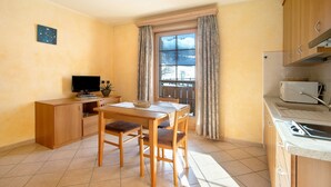 A compact kitchen with a dining area, wooden cabinets, a window with curtains, and a flat-screen TV.