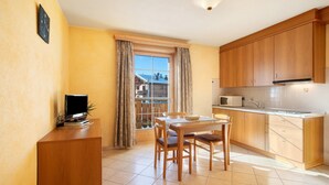 A compact kitchen with wooden cabinets, a dining table with chairs, a small TV on a cabinet, and a view through a window.