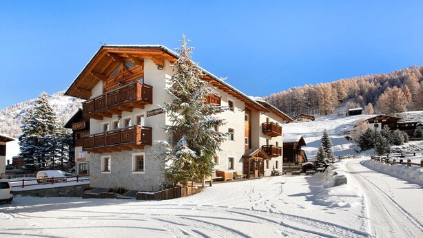 A snow-covered building with wooden balconies, a ski slope in the background, and a clear blue sky.