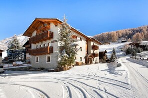 Un bâtiment enneigé avec des balcons en bois, une piste de ski en arrière-plan et un ciel bleu dégagé.