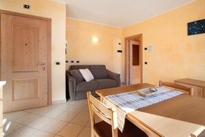 A room with a wooden door, a sofa, a dining table with a placemat, and a wall clock. - Apartment 'Al Bait Da Marangona 3' with Mountain View, Shared Garden and Wi-Fi (Livigno)