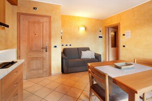 Ein kleines Zimmer mit Holztür, einer Couch, einem Esstisch mit Stühlen und Fliesenboden.