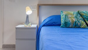 A bed with a blue mattress and a pillow with a tropical leaf pattern, a bedside lamp, and a small drawer unit. - Apartment 'Brezza Marina In Oasi Del Mare' with Sea View and Wi-Fi (Granelli)