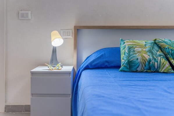 A bed with a blue mattress and a pillow with a tropical leaf pattern, a bedside lamp, and a small drawer unit.