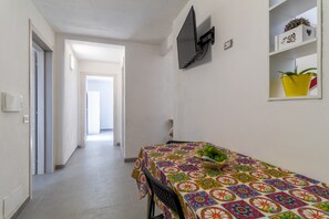 Un couloir mène à une salle à manger où une table est dressée pour deux personnes. On y trouve également un téléviseur fixé au mur et une étagère ornée d’objets décoratifs.