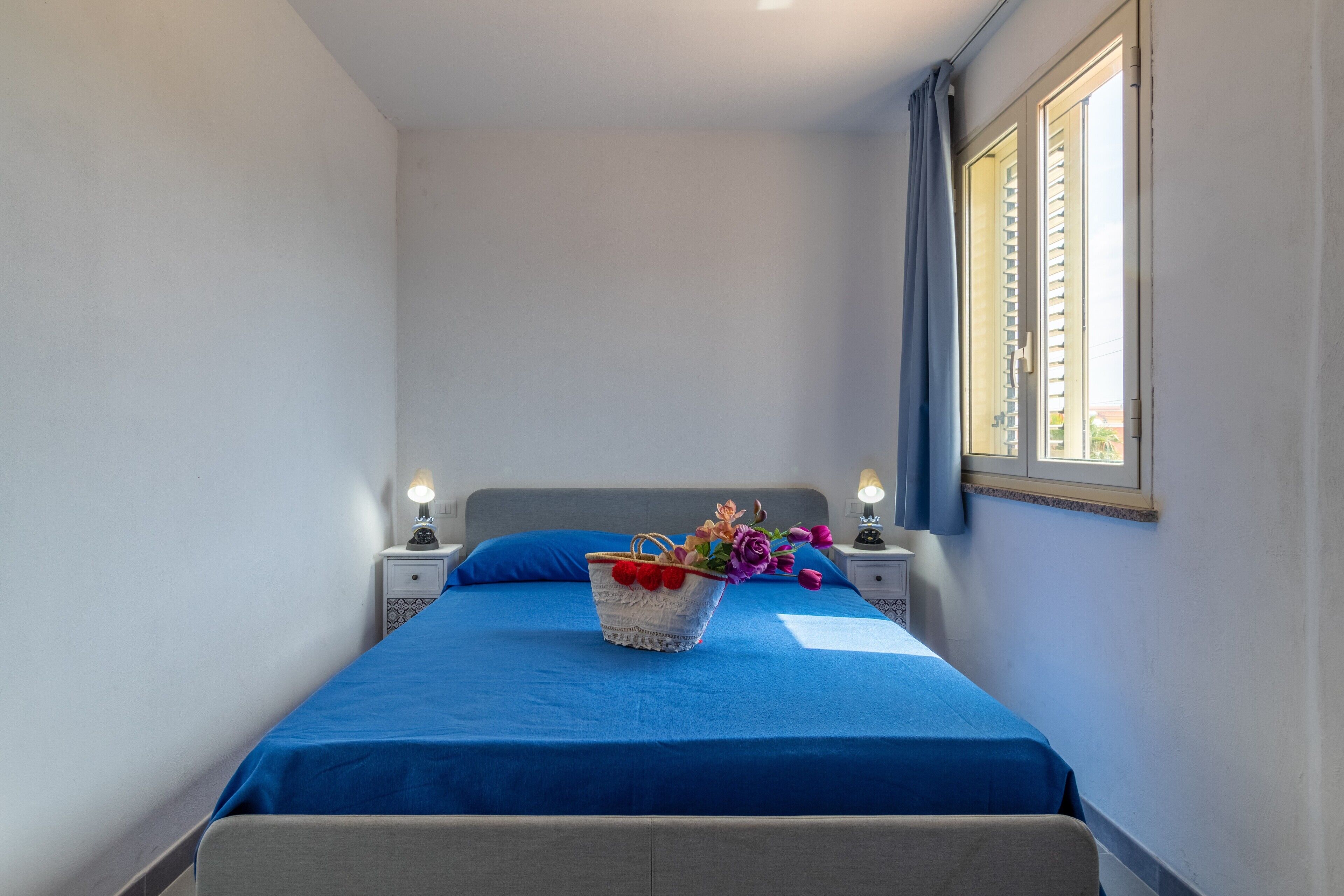 Een slaapkamer met een bed, nachtkastje, lamp, raam met gordijnen en een vaas met bloemen.