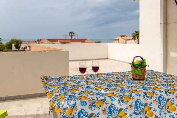 Une table recouverte d’une nappe à motifs floraux, deux verres de vin rouge et un panier de citrons, le tout sur un balcon donnant sur une zone côtière.