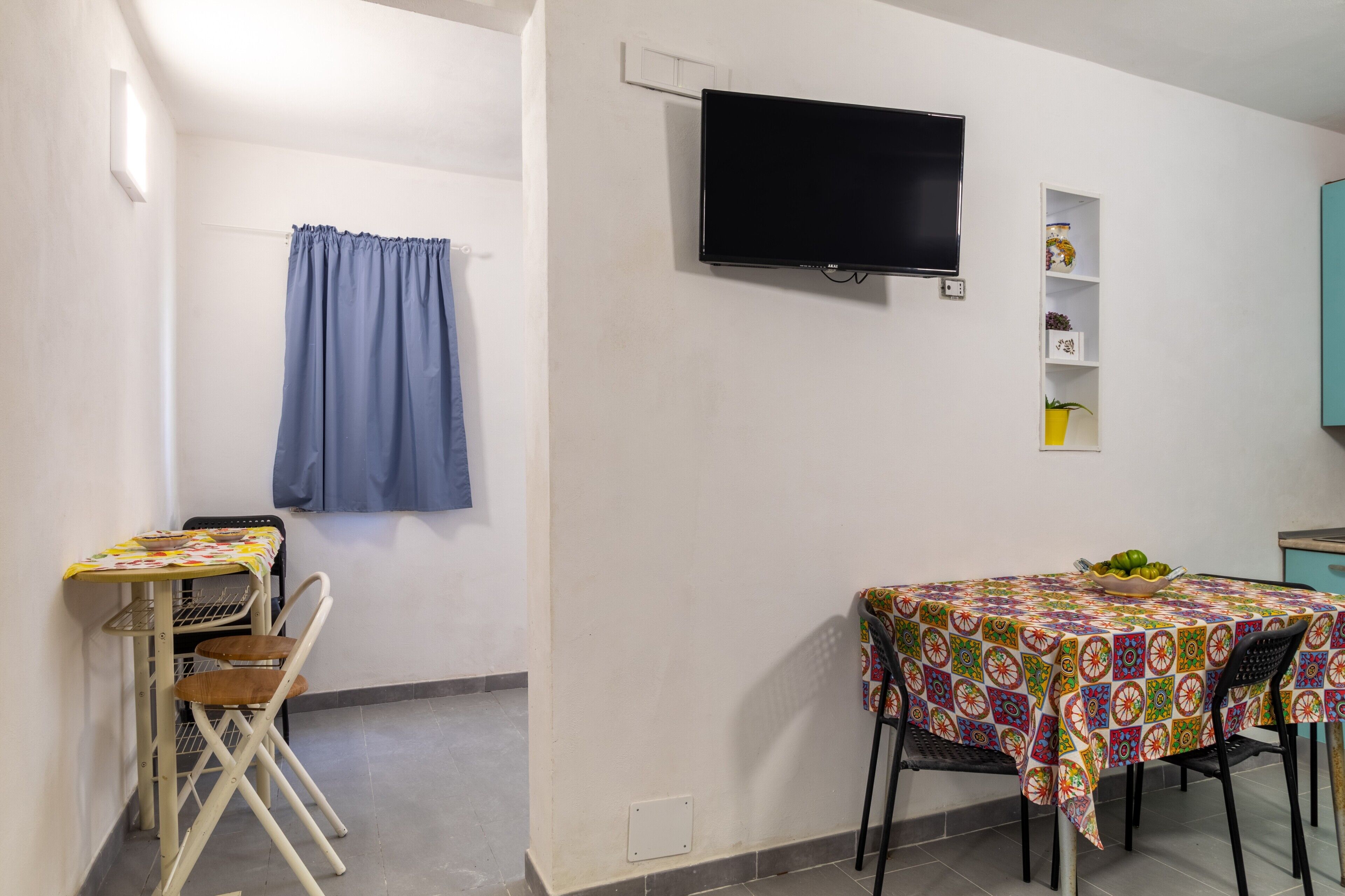 A room with a TV mounted on the wall, a table with chairs, a shelf with decorative items, and a curtain.