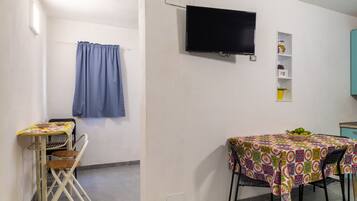 A room with a TV mounted on the wall, a table with chairs, a shelf with decorative items, and a curtain.