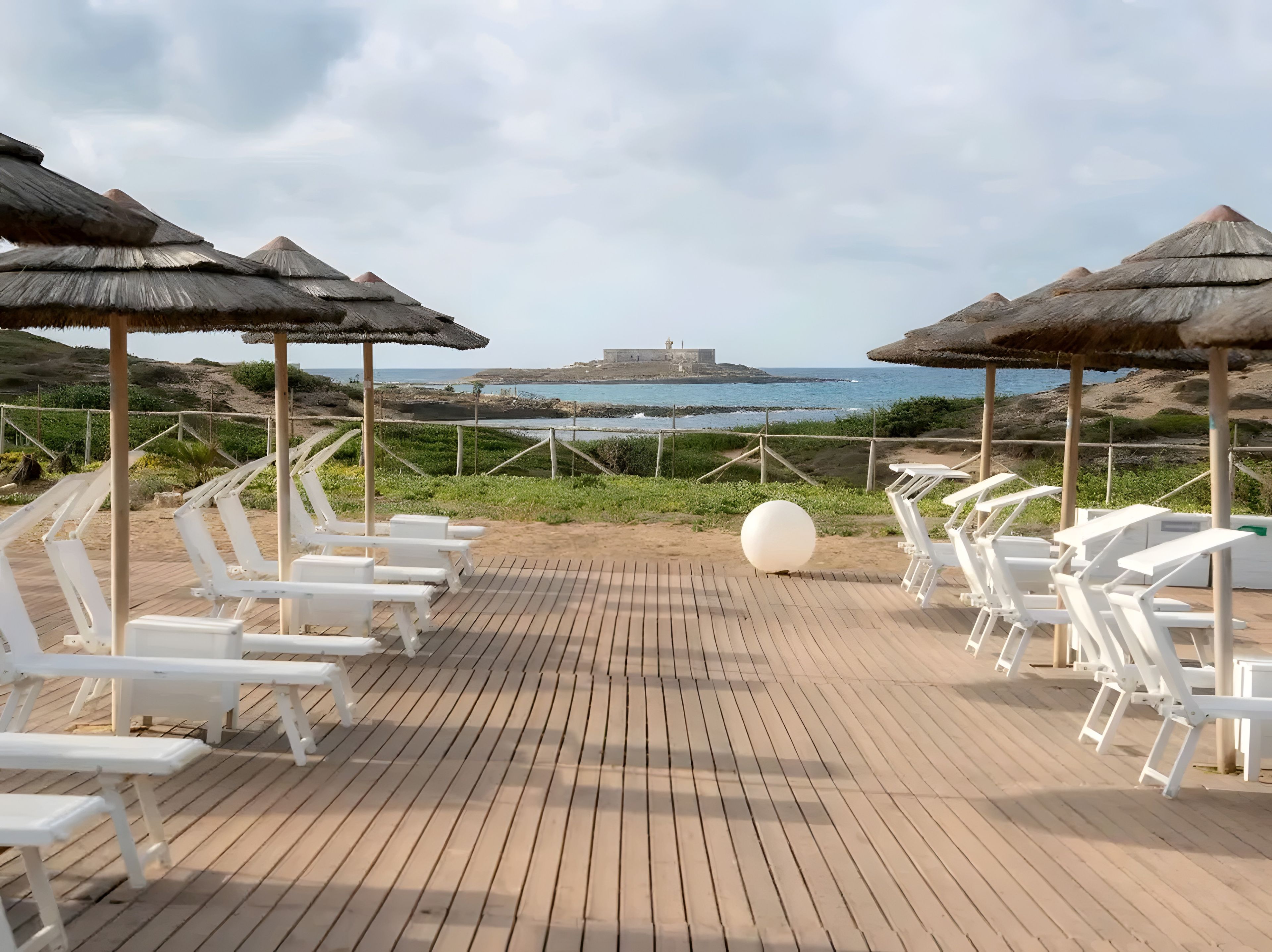 A wooden deck with white lounge chairs and thatched umbrellas, overlooking a rocky coastline and the sea.