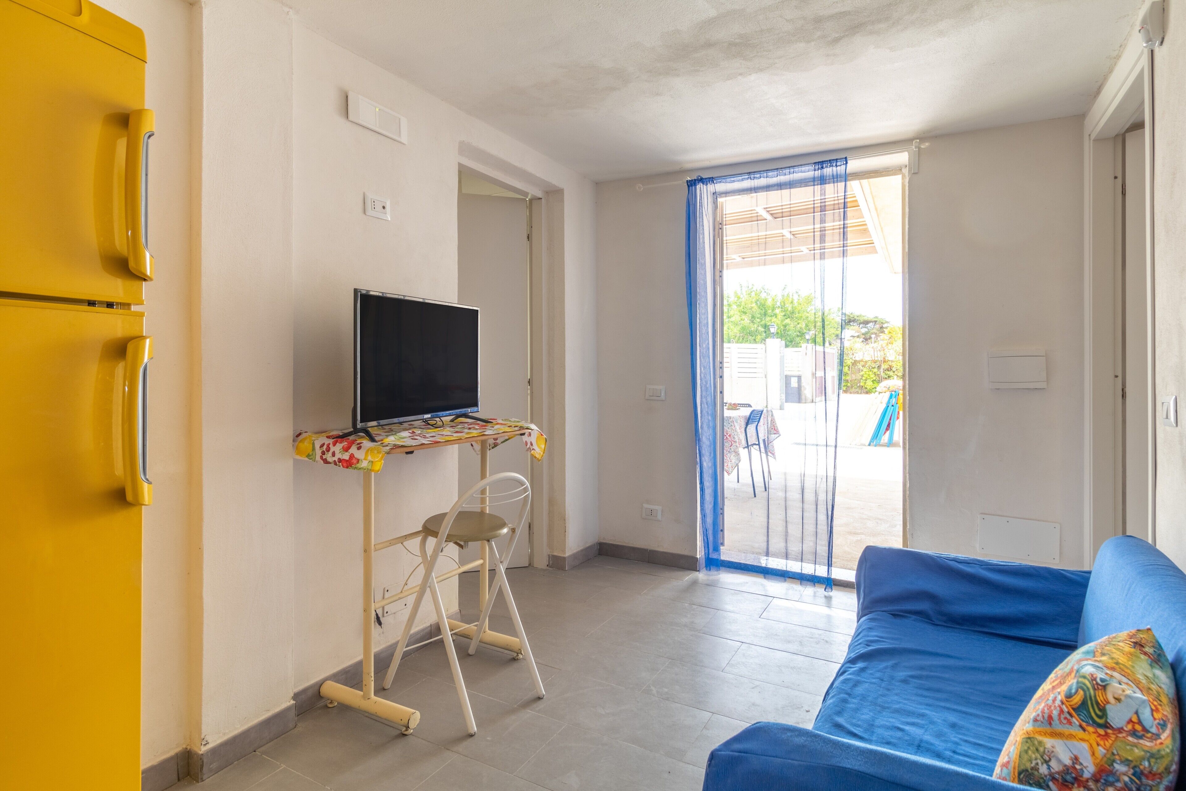 Ein Zimmer mit einem blauen Sofa, einem Fernseher auf einem kleinen Tisch, einem gelben Schrank und Blick durch eine Glastür.