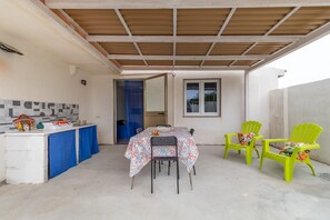 Ein überdachter Außenbereich mit Tisch und Stühlen, einer kleinen Küchenzeile und einer Tür zu einem weiteren Raum.