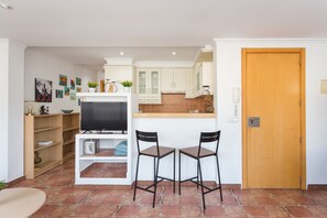 Un espace de vie compact comprenant un coin cuisine, un téléviseur, deux tabourets de bar et une porte en bois.