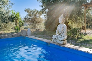 Un entorno exterior tranquilo con una estatua junto a una piscina, rodeado de árboles y una pérgola.