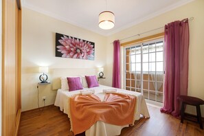 A bedroom with a bed, a large window with curtains, a painting on the wall, and a ceiling light.