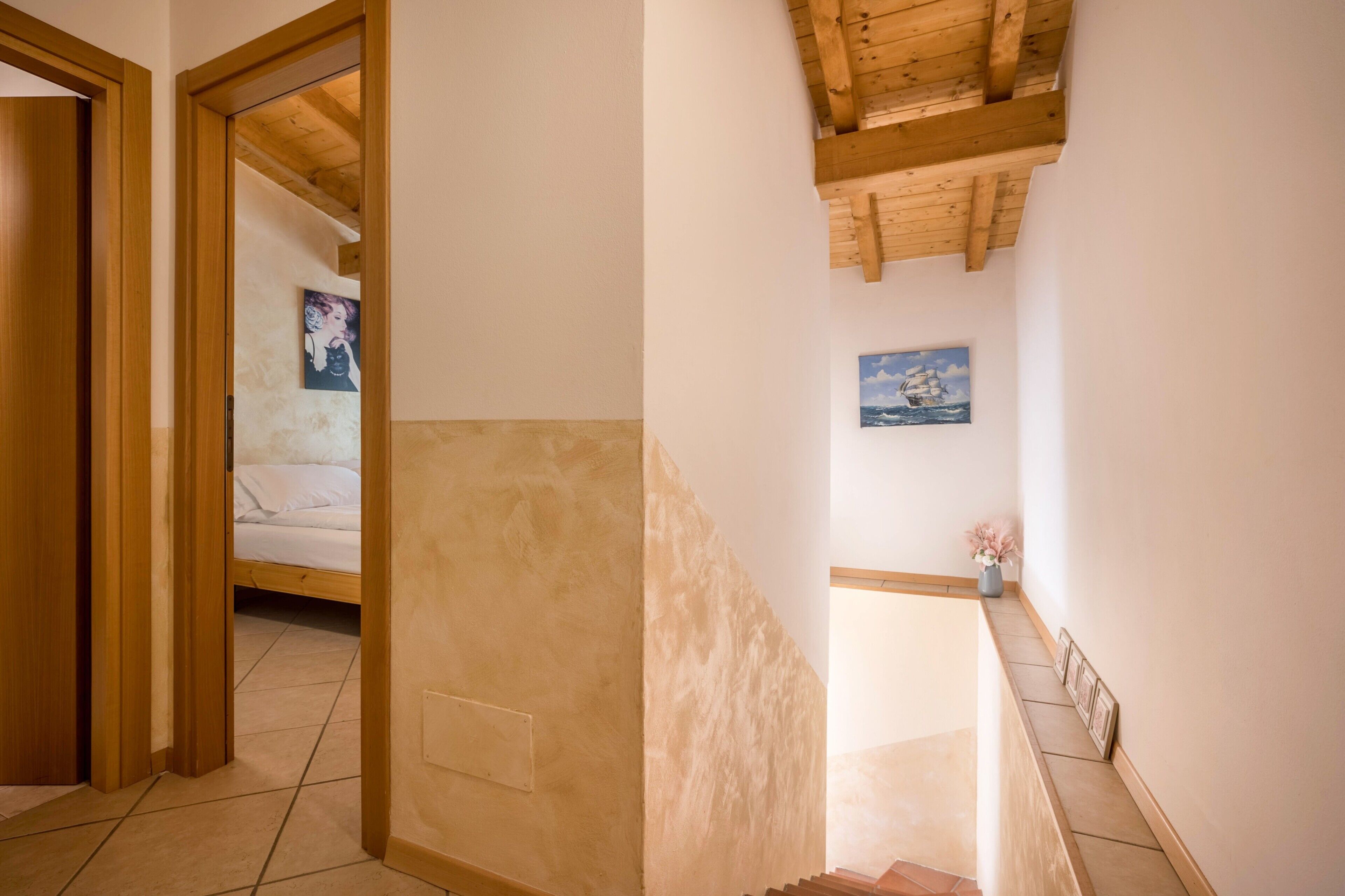 A hallway with a wooden door, a bed with white bedding, a painting on the wall, and a small vase with flowers.