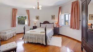 A bedroom with a wooden floor, a bed with a white bedspread, two bedside tables with lamps, a bench, and a large window with curtains. - Holiday Home 'Moinho De Paradela' with Private Pool, Private Terrace and Wi-Fi (Paradela)