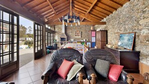 Une pièce spacieuse avec un plafond haut, des murs en pierre et de grandes fenêtres. On y trouve deux fauteuils en cuir avec des coussins, une table en bois et une cheminée en pierre.