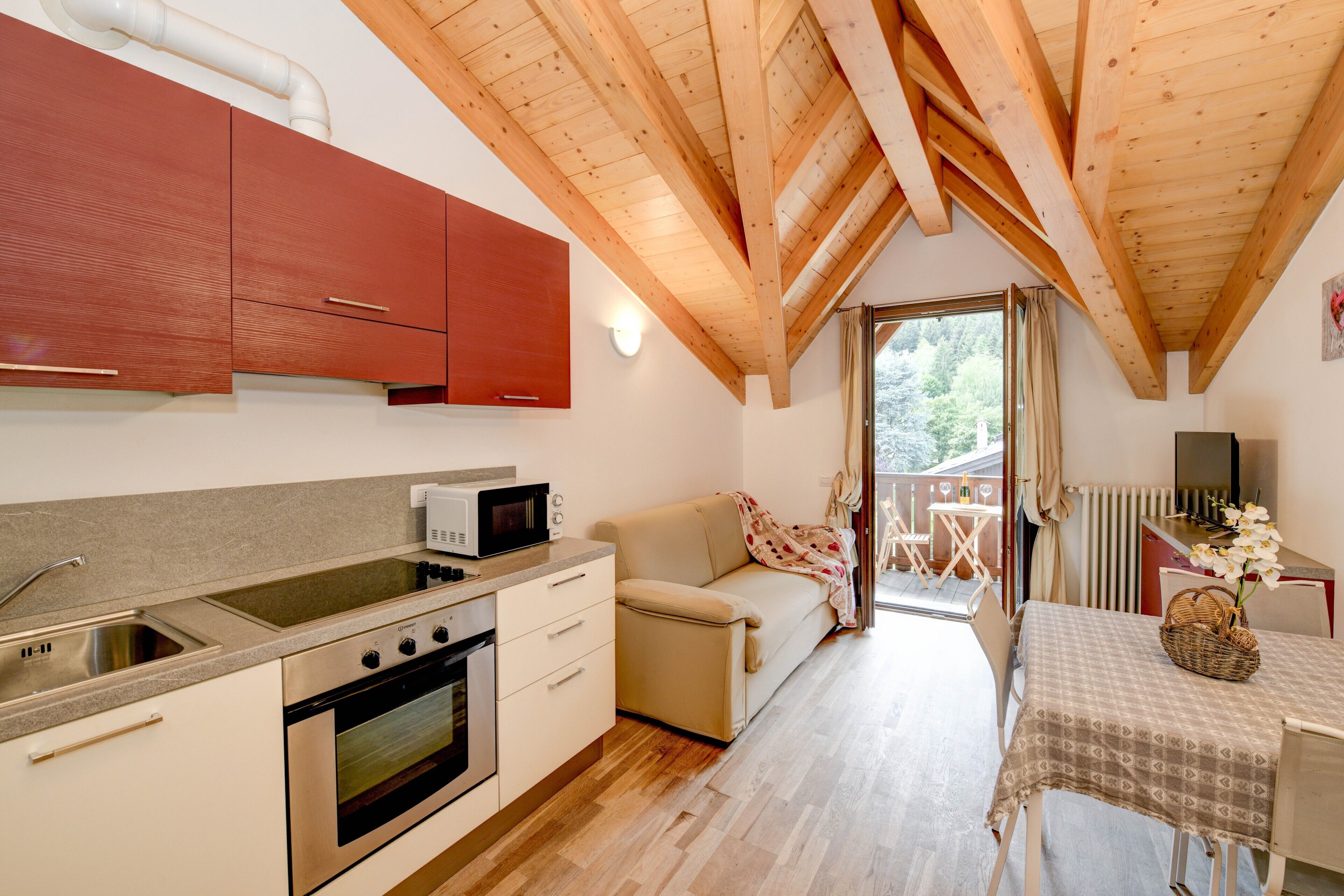 A compact kitchen with a microwave, stove, and sink, a sofa, a dining table with a basket, and a view through a glass door to an outdoor area.