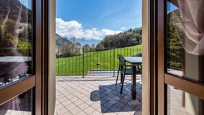 Un balcon avec une table et des chaises, une vue sur un champ verdoyant et des montagnes à l’horizon.