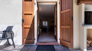 Une porte en bois donnant sur un intérieur carrelé, un paillasson noir à l’entrée et une chaise à l’extérieur.