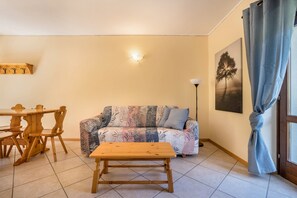 A living room with a sofa, a wooden table, a dining table with chairs, a lamp, and a painting on the wall.