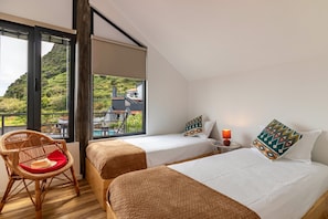 Une chambre avec deux lits, un fauteuil en osier, une petite table avec une lampe et une vue sur un paysage montagneux.