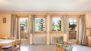 Una habitación con una mesa de comedor de madera, sillas, un sillón con cojín y ventanas grandes con cortinas.