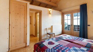 Quarto com vigas de madeira, cama com colcha xadrez, uma cadeira e janela com cortinas.