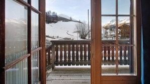 Uma varanda de madeira com guarda-corrimão de metal, vista para uma paisagem montanhosa coberta de neve e céu claro.
