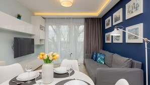 Una sala de estar moderna con comedor, que incluye un sofá gris, un televisor de pantalla plana, una mesa de comedor con platos y vasos, y un jarrón con flores amarillas.