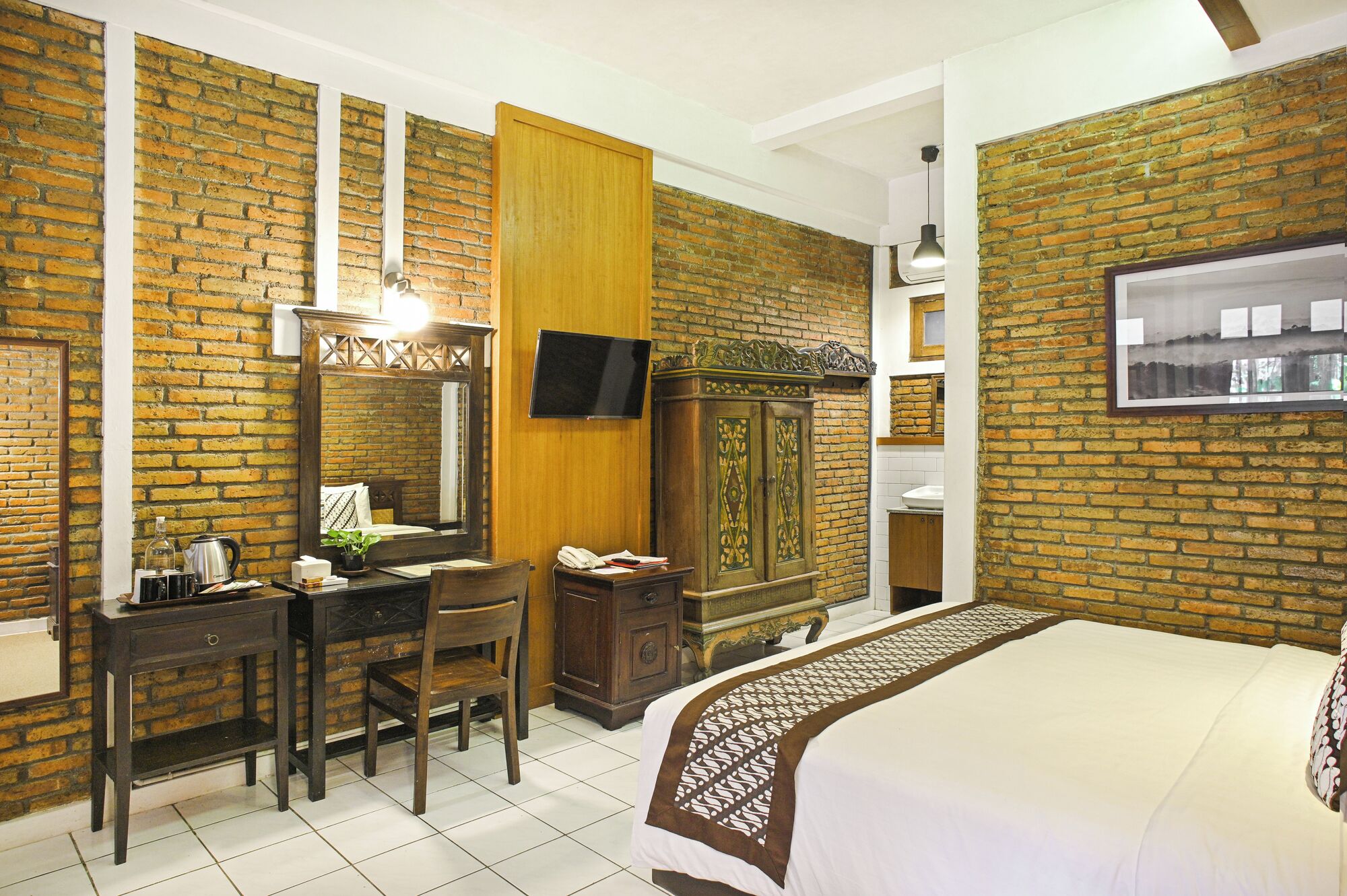 Une chambre d’hôtel avec un mur en briques, une armoire en bois, un bureau avec une chaise, un lit recouvert d’une couvre-lit à motifs et un téléviseur fixé au mur.