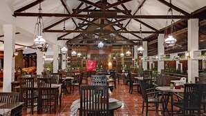 Un restaurante espacioso con techos altos y vigas a la vista, que cuenta con mesas y sillas de madera, lámparas colgantes y plantas en macetas.