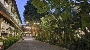 Un sendero adoquinado bordeado de plantas en macetas y luces colgantes, que conduce a un edificio con fachada de madera y vistas a un jardín exuberante.