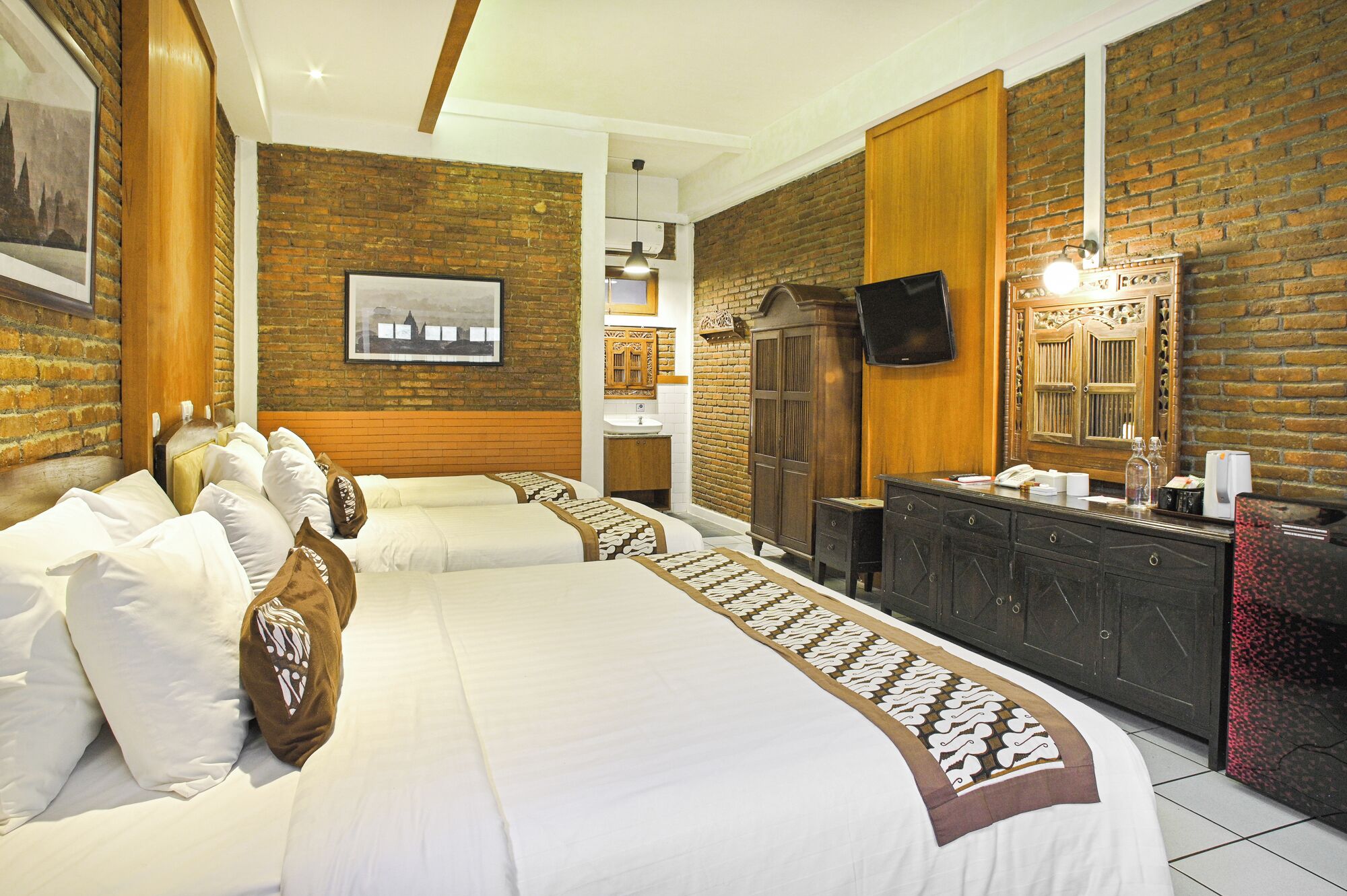 A hotel room with two beds, a wooden headboard, a brick wall, a mounted TV, a wooden cabinet, and a bathroom with a sink and mirror.