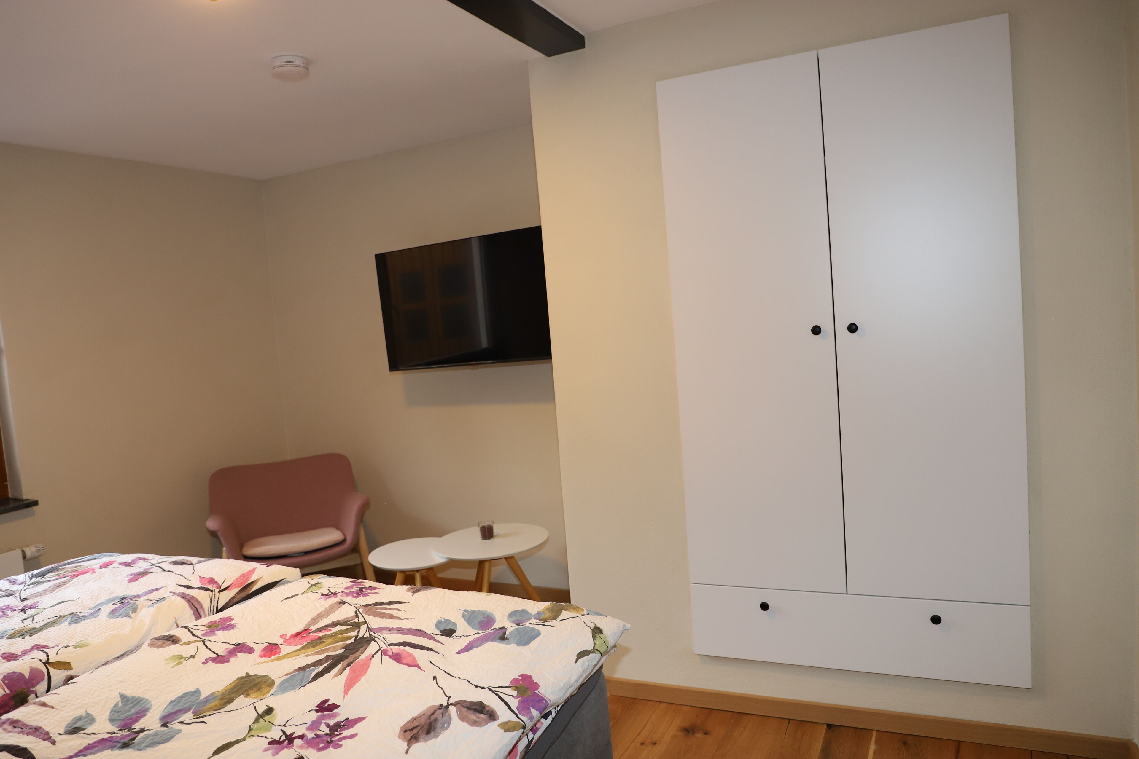 Une chambre à coucher avec un lit, une armoire, une chaise rose, une petite table ronde et un téléviseur fixé au mur.