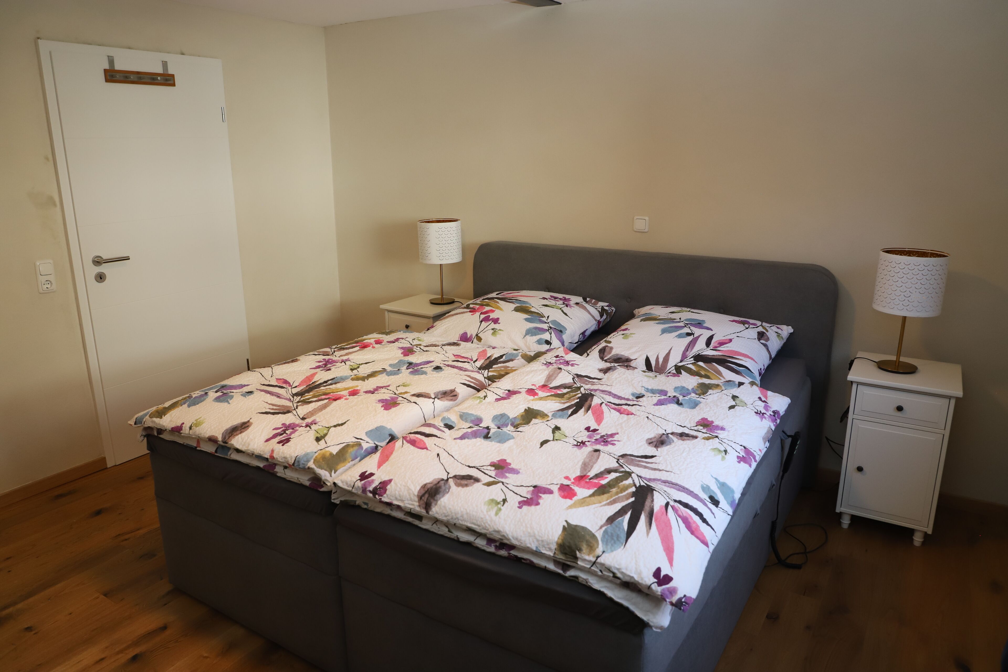 A bedroom with a bed, two bedside tables, a nightstand, a door, and a wall socket.