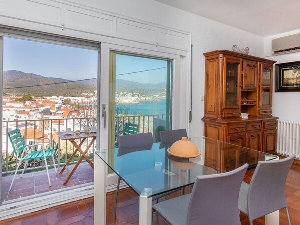 Un balcón con una mesa y sillas de vidrio, un armario de madera y vistas a un pueblo costero.