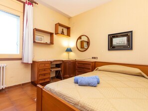 Un dormitorio con cama, escritorio, silla, lámpara y un cuadro en la pared.