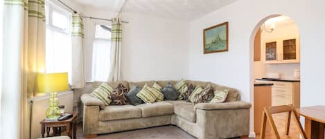 A cozy living room with a sectional sofa, a small table with a lamp, a chair, and a kitchen area with cabinets and a countertop.