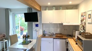 Une cuisine moderne avec des armoires blanches, un comptoir en bois et un four encastré. On y trouve un coin repas avec une table et des chaises, ainsi qu’un téléviseur fixé au mur.