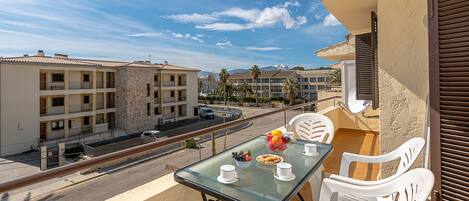 Un balcon avec une table en verre préparée pour le petit-déjeuner, des chaises en plastique blanches et une vue sur les bâtiments voisins et une route.
