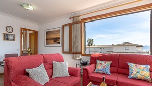 A living room with two red sofas, a coffee table with a bottle and glasses, a framed picture, and a view of buildings through a window. - Apartment LLenaire in Puerto Pollensa by Home Villas 360 (Puerto Pollensa)