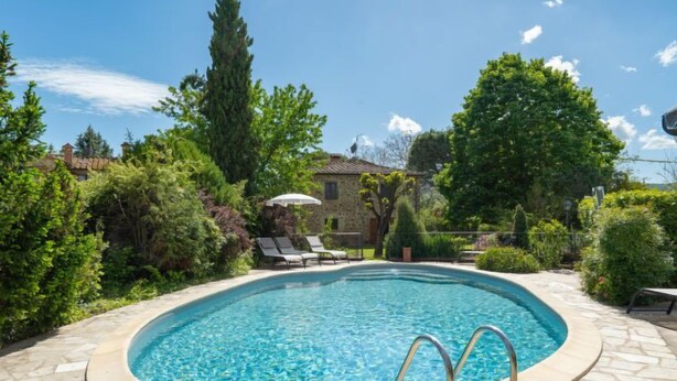 A swimming pool with a stone deck, surrounded by lush greenery and a stone building in the background.